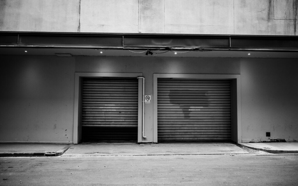 A monochrome urban exterior with two closed garage doors and a concrete wall.